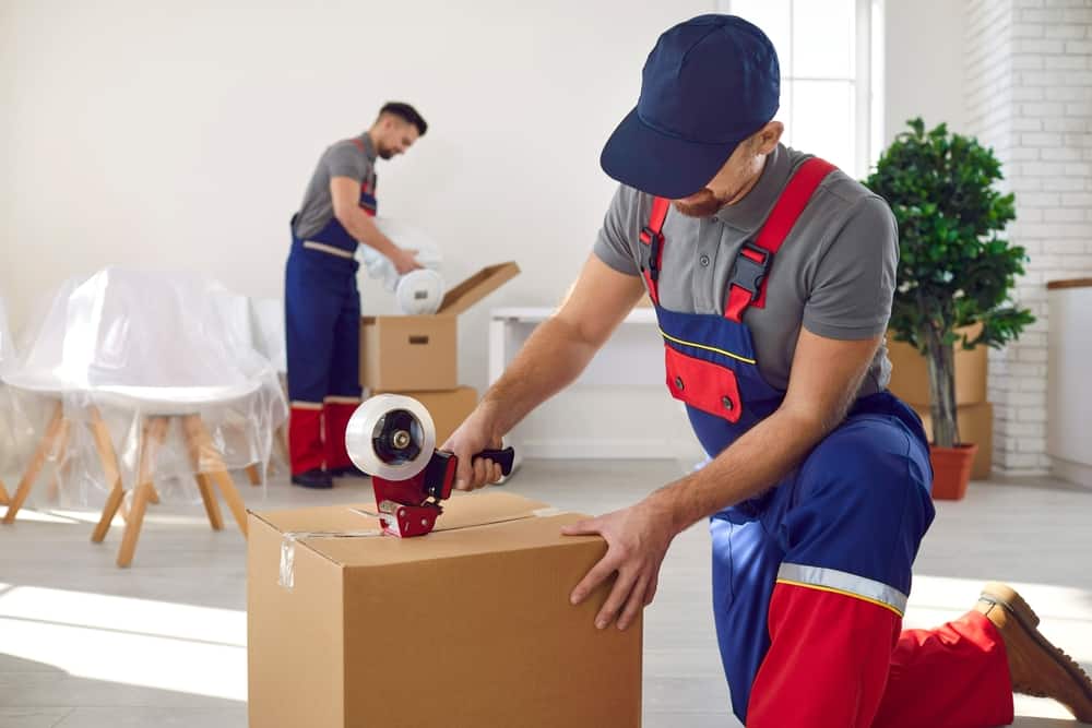 Young worker in uniform holding packing duct tape gun roller dispenser and sealing fragile cardboard box with sticky adhesive tape. Moving house, relocation company and truck delivery services concept